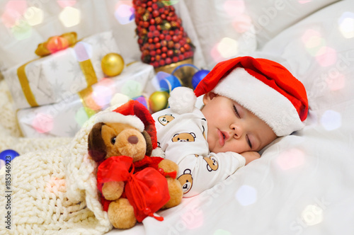 Little baby boy sleeping in pajamas and Santa hat with a toy dog and a gift in a red bag on the background of gift boxes
