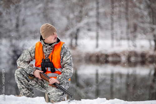 Male hunter in camouflage, armed with a rifle, sittiing in a snowy winter forest