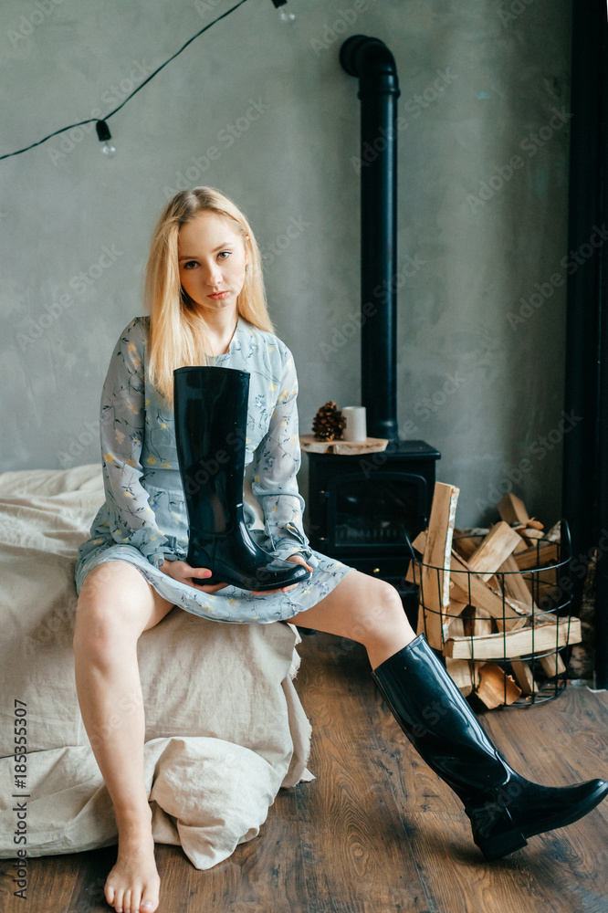 Beautiful young blonde girl posing for camera. Odd strange crazy bizarre  unusual female sitting on sofa in loft style room. Funny barefoot woman  holding black boot. Stylish fashion woman portrait. Stock Photo