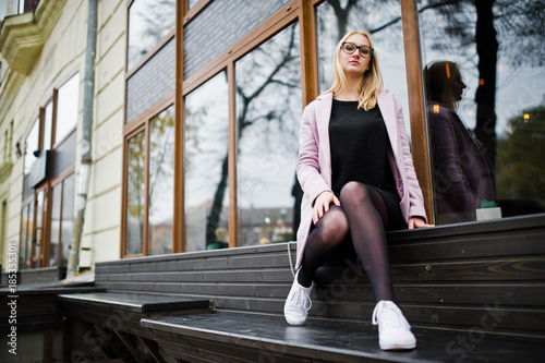 Blonde girl at glasses and pink coat, black tunic sitting on bench at street.