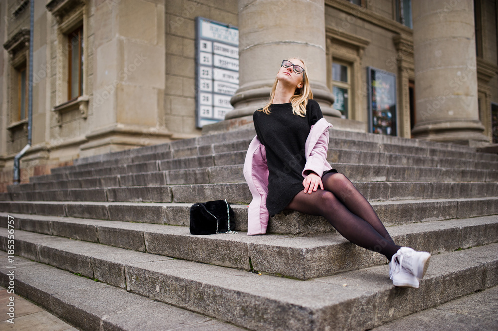 Blonde girl at glasses and pink coat with handbag sitting on stairs of opera building. Blonde girl at glasses and pink coat with handbag sitting on stairs of opera building.