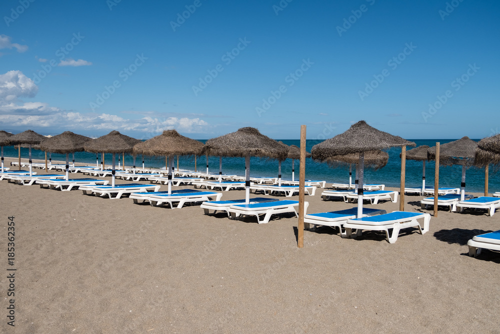 Line of straw beach umbrellas and sun loungers with clear blue sea and sky in background