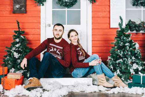 Happy beautiful young couple in love dressed in sweater sitting on porch steps at light house with decorated in red green New Year door at home. Christmas or birthday pipes. Family and holiday 2018. © speed300