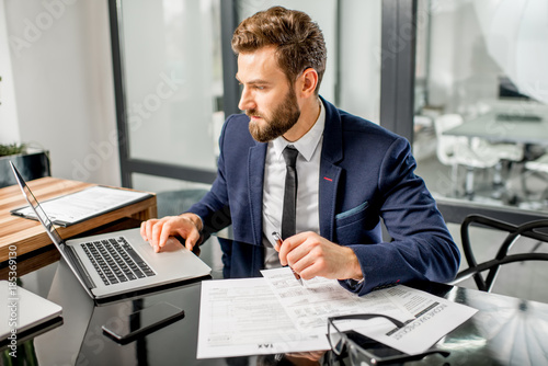 Handsome tax manager dressed in the suit working with documents and laptop at the modern office interior photo