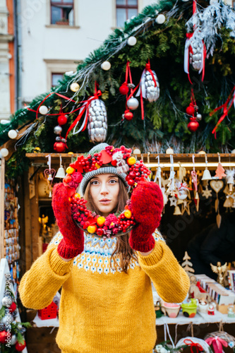 Vertical portrait of surprised positive hipster woman looking through Christmas wreath over kiosk outdoors at christmas fair market on central square of the european city.