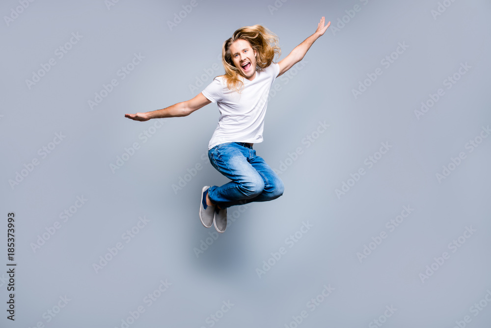 Handsome carefree young delightful guy with long blonde hair is screaming and jumping up, isolated on grey background