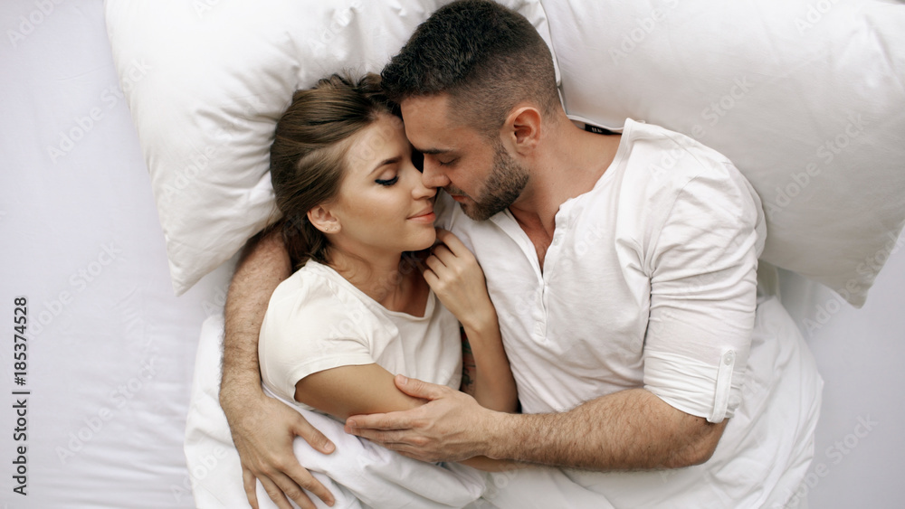 Young beautiful and loving couple kiss and hug into bed while waking up in the morning. Top view of attractive man kissing and talking his smiling wife Young beautiful and loving couple kiss and hug into bed while waking up in the morning. Top view of attractive man kissing and talking his smiling wife