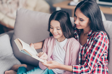 It's so interesting and exciting! Young smiling mother is reading a funny book for her cute sweet little daughter, they are doing homework and embracing and spending time together