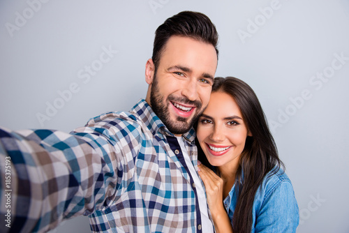 Self portrait of young, caucasian, attractive, lovely, cute, cheerful, positive, couple in shirts, man making selfie with his wonderful woman on mobile phone over grey background