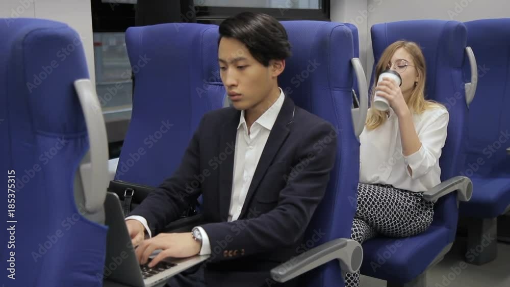 The asian businessman and blond female travel by train and sit one after another. The man in black siut and with stylish haircut works on his laptop and the woman in white blouse and round glasses The asian businessman and blond female travel by train and sit one after another. The man in black siut and with stylish haircut works on his laptop and the woman in white blouse and round glasses