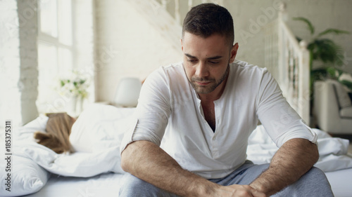 Upset young man sitting in bed suffer of problems while his girlfriend sleep in bedroom