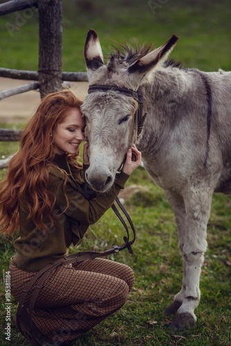 A girl with curly red hair in fashionable clothes in the style of Provence hugs a cute donkey © selenit