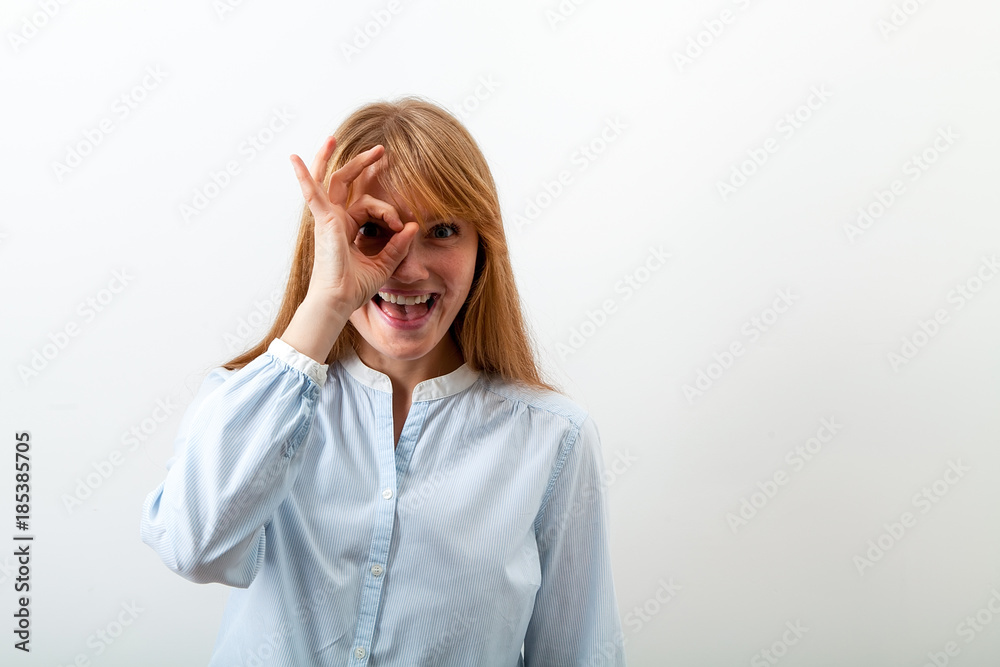 Studio indoor portrait of red-headed girl with freckles dressed in casual blue shirt showing a sign of approval with her fingers and smiling happily. White background, copy space. Studio indoor portrait of red-headed girl with freckles dressed in casual blue shirt showing a sign of approval with her fingers and smiling happily. White background, copy space.