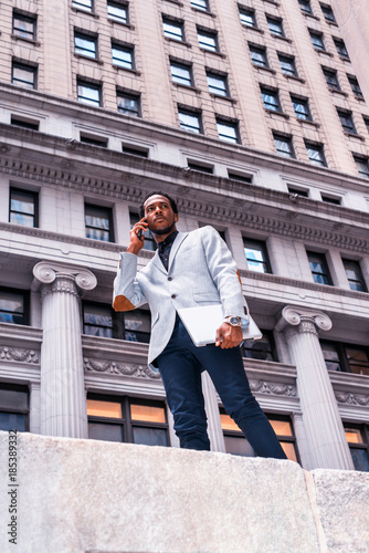 Power of Technology. African American businessman working in New York, wearing gray blazer, wristwatch, standing on street, holding laptop computer, calling on mobile phone. Photo stylized look..