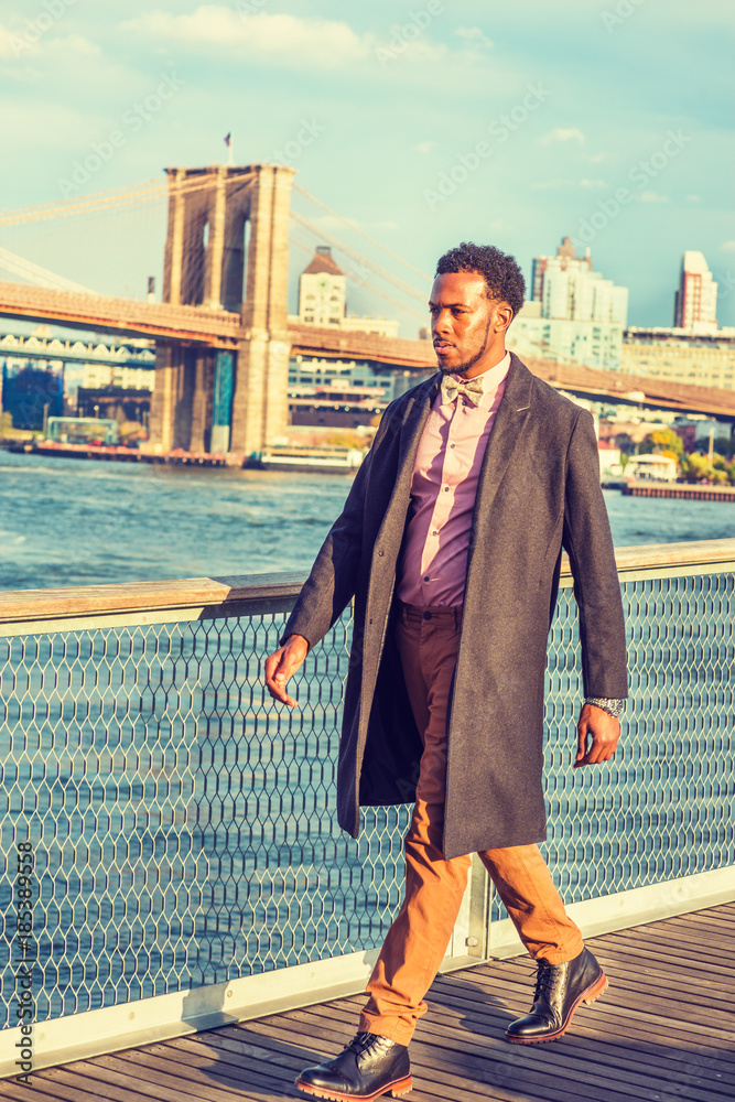 African American Businessman Traveling In New York. Wearing Woolen  Overcoat, Pink Shirt, Brown Pants, Leather Boot Shoes, Bow Tie, Black Man  With Beard, Confidently Walking On Deck At Harbor In Sunset Stock