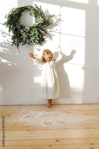 Playful little blonde female child in white dress, throws artificial snow in air, poses in white cozy room with symbolic Christmas garland on wall, enjoys winter holidays. Children, emotions concept photo