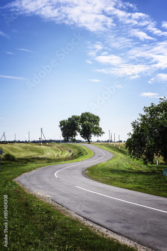 a curving country road with an old tree hedgerows and meadows under a blue cloudy summer sky in Belarus photo