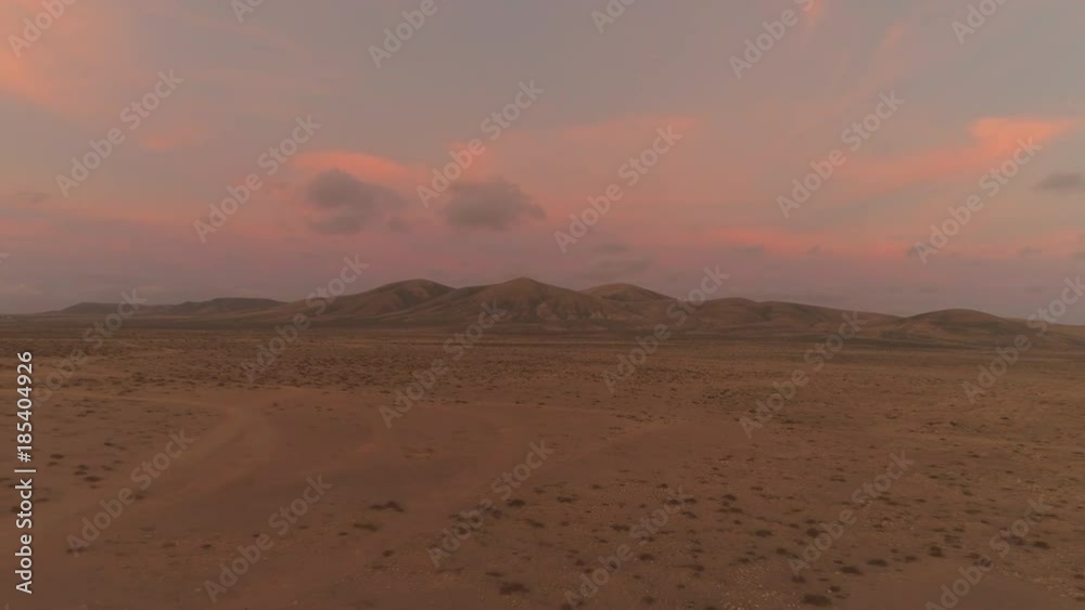 AERIAL: Flying away from big volcanic structure in pink colored summer sunset in lonely desert. Empty sand roads leading through dried up nature. Volcanic structure towering over the lonely wilderness