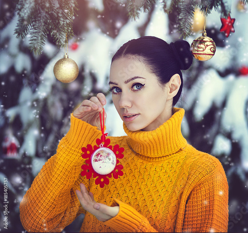A beautiful girl with blue eyes in a yellow sweater is holding a New Year's toy under a snow-covered tree. A girl in fairy-tale New Year's decorations. Magic winter forest. photo