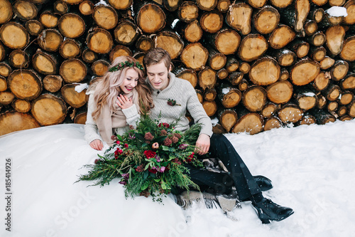 Bride and groom tenderly looks at each other. Stylish newlyweds sits on snow on the wooden background. Winter wedding photo