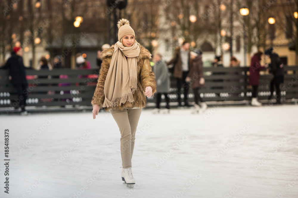 Full length portrait of young female with blonde hair in  fur coat, beige hat, scarf and trousers skating on ice rink, outdoors at winter / Weekends activities outdoor in cold weather/