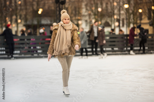 Full length portrait of young female with blonde hair in fur coat, beige hat, scarf and trousers skating on ice rink, outdoors at winter / Weekends activities outdoor in cold weather/