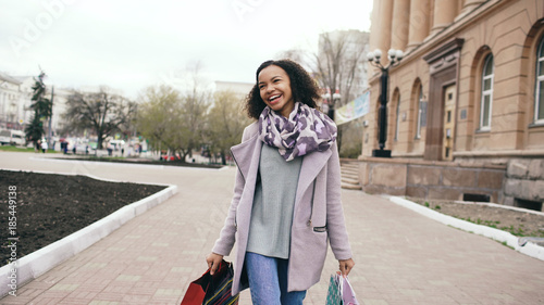 Attractive mixed race girl dancing and have fun while walking down the street with bags. Happy young woman walking after shopping on mall sale