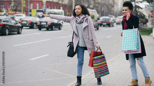 Two attractive african american women with shopping bags calling for taxi cab while coming back from mall sales