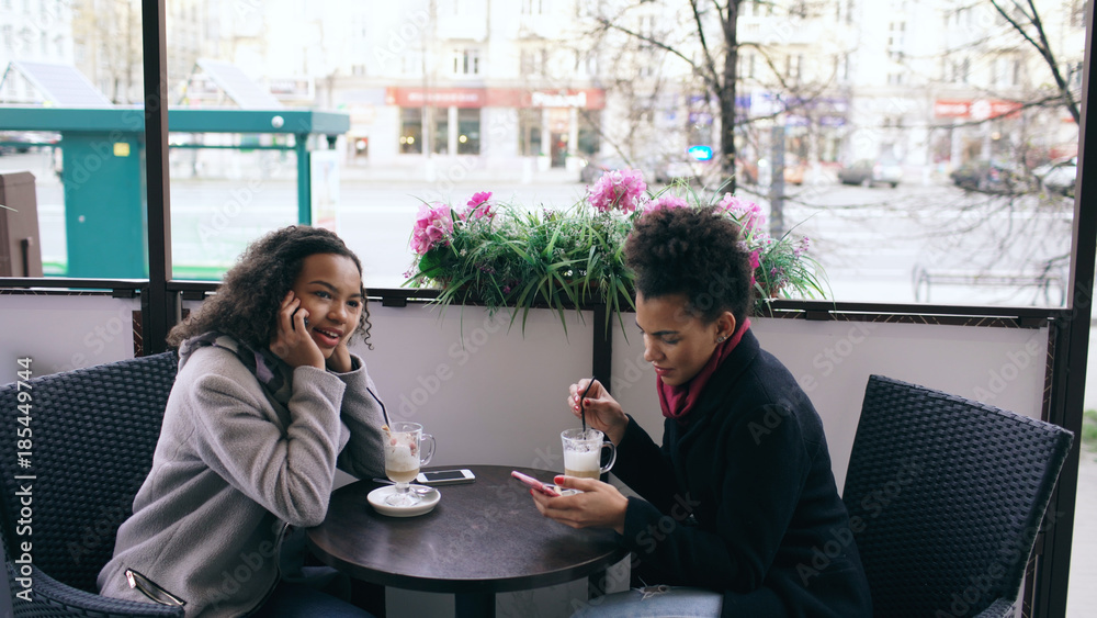 Attrcative mixed race woman sitting at the table in the street cafe talking cell phone while her friend surfing smartphone Attrcative mixed race woman sitting at the table in the street cafe talking cell phone while her friend surfing smartphone