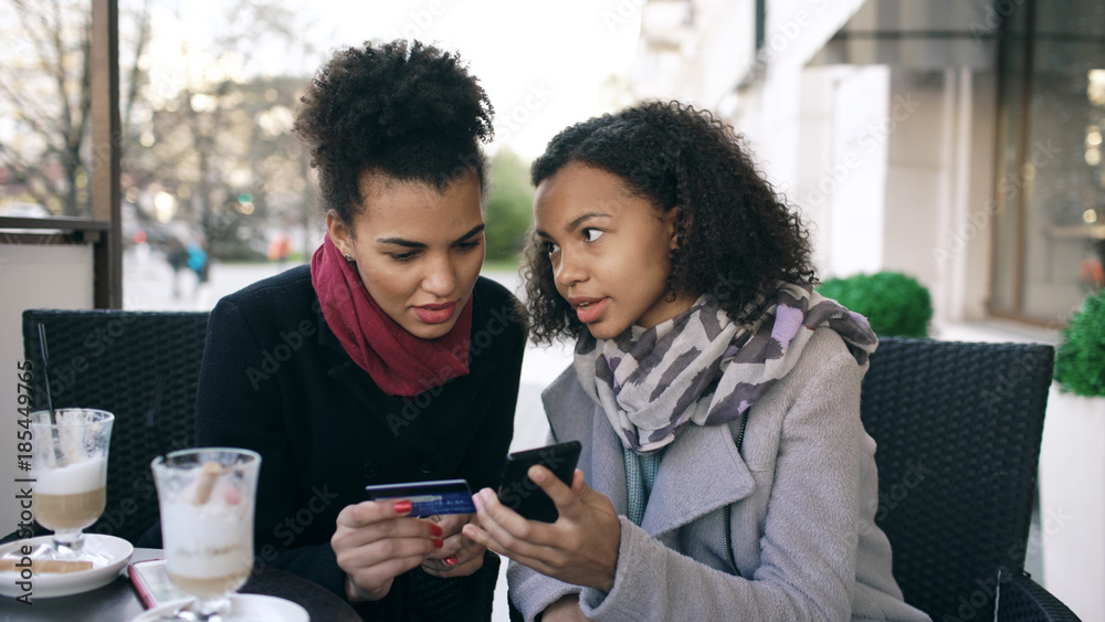 Fototapeta premium Two attractive mixed race women having online shopping with credit card and smartphone while talking and drinking coffee in street cafe