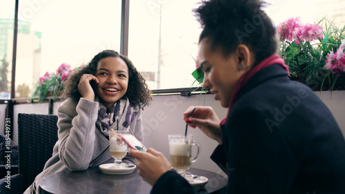 Two mixed race women sitting at the table in the street cafe using smartphone and talking