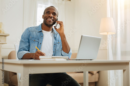 Speaking. Handsome gleeful dark-haired afro-american businessman smiling and talking over the phone and holding a pencil while sitting at the table and working on his laptop