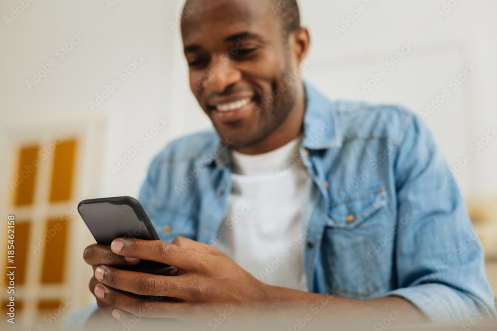 Phone. Handsome gleeful young afro-american man smiling and writing a message on his phone and looking at the screen Phone. Handsome gleeful young afro-american man smiling and writing a message on his phone and looking at the screen