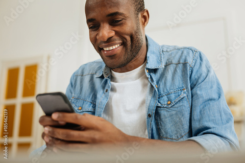 Good mood. Handsome alert young afro-american man smiling and writing a message on his phone and looking at the screen