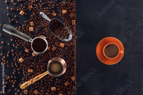top view of roasted coffee beans, scoop, coffee pot, coffee tamper and cup of coffee on black