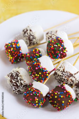 marshmallows on a sticks, laying on a white plate. glazed with chocolate and colorful sprinkles.