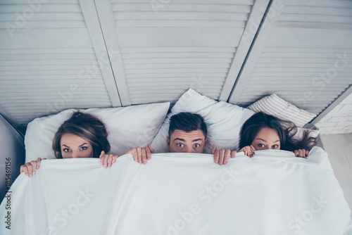 Top view of funny three partners hiding behind blanket, peeking over sheets and looking at camera while lying in bed at home, weekend, rest, relax, someone catches them photo