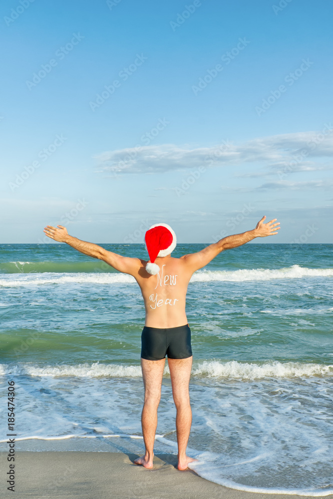 Man in Santa hats with the inscription New Year on the back on the sea coast. Hands up. Back view Man in Santa hats with the inscription New Year on the back on the sea coast. Hands up. Back view