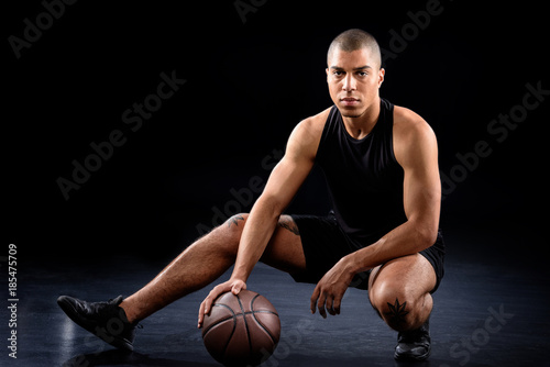 handsome african american basketball player sitting with ball on black