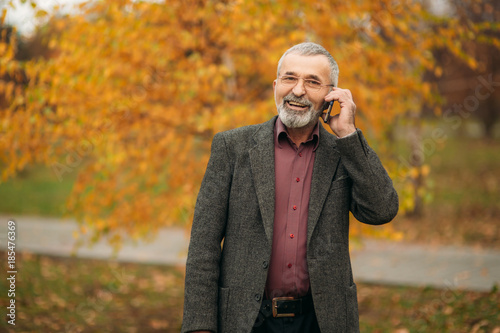 A handsome elderly man wearing glasses is using a phone. Walk in the park in autumn © Aleksandr