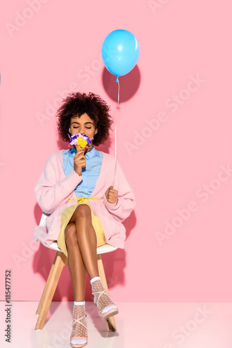 african american woman with flowers in ice cream cone and balloon in hands on pink wall backdrop
