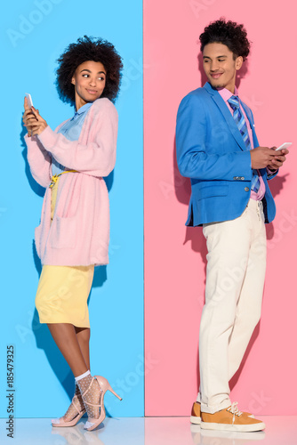 Young african amercian smiling couple with phones standing back to back on pink and blue background