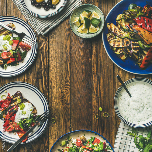 Flat-lay of healthy dinner table setting. Fresh salad, grilled vegetables with yogurt and dill sauce, pickled olives, lemon water over wooden background, copy space, square crop. Clean eating photo