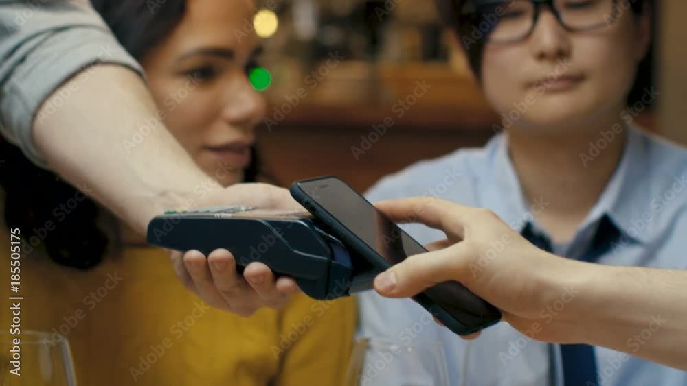 In the Bar Close-up Of Waiter's Extended Hand with Card Machnine and Man Paying with Contactless Mobile Phone. In the Background Group of Diverse Young People Having Fun.