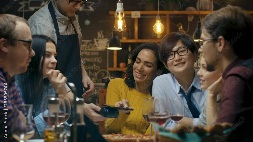 In the Bar Waiter Holds Credit Card Payment Machine and Beautiful Woman Pays for Her Order with Contactless Mobile Phone Payments System. She's Surrounded by Dear Friends and Has Time of Her Life. 