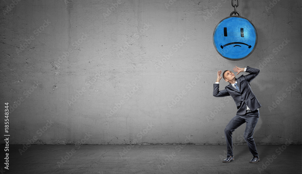 Obraz premium A scared businessman stands on concrete background under a wrecking ball with a painted blue sad face.