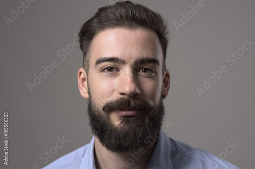Close up portrait of young elegant bearded adult man with brown hair and eyes looking at camera over gray studio background. 