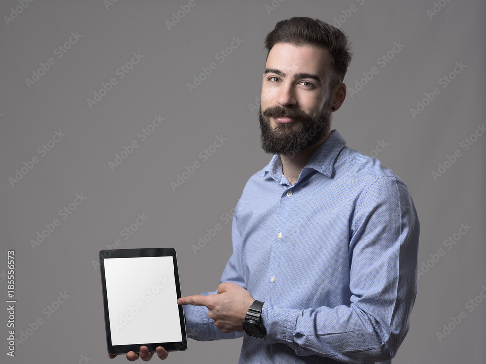 Young smiling friendly business man pointing finger and advertising empty blank tablet computer monitor over gray studio background. Young smiling friendly business man pointing finger and advertising empty blank tablet computer monitor over gray studio background.