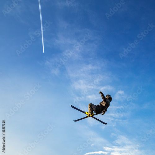 Ski jumper flying high in the air on a blue sky background with a trace of a jet aircraft.