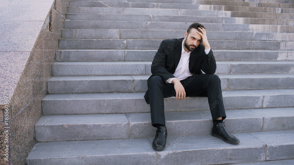 Upset young business man having stress and sitting on stairs in street. Businessman having deal problems concept Upset young business man having stress and sitting on stairs in street. Businessman having deal problems concept